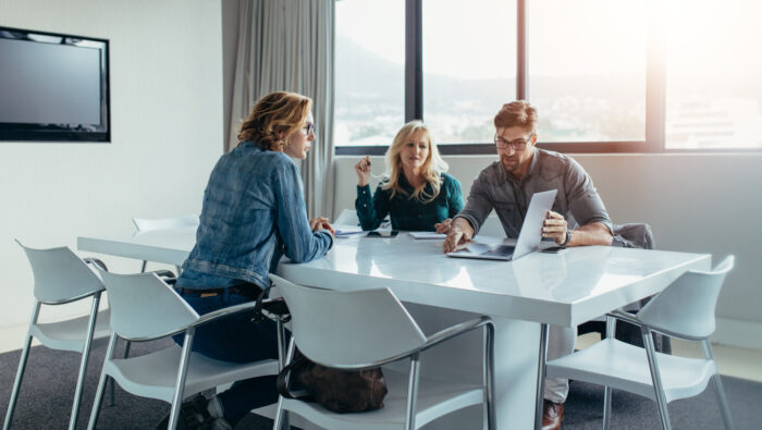 Trois personnes sont assises à une table blanche dans une salle de réunion lumineuse. Un conseiller en construction Blavier montre quelque chose sur un ordinateur portable aux autres. Ils sont engagés dans une discussion enthousiaste, avec de grandes fenêtres en arrière-plan laissant entrer la lumière naturelle.
