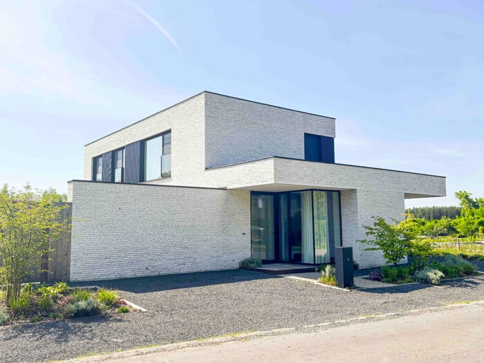 Une maison moderne à deux étages avec une façade en briques blanches, de grandes fenêtres et un toit plat. L'entrée principale est dotée de portes vitrées et le jardin est aménagé de façon minimaliste avec de petits arbustes et du gravier.
