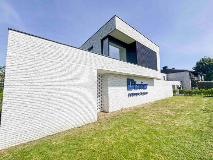 Un bâtiment moderne en briques blanches, doté de grandes fenêtres et d'un toit plat, trône sur une pelouse. Sur le mur face à la caméra, une pancarte « Denier Ramen en deuren » est affichée. Le ciel est clair et bleu.