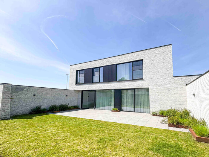 Maison moderne à deux étages avec un extérieur en briques blanches, de grandes fenêtres à cadre noir et un patio spacieux donnant sur une pelouse verte, sur fond de ciel bleu clair.