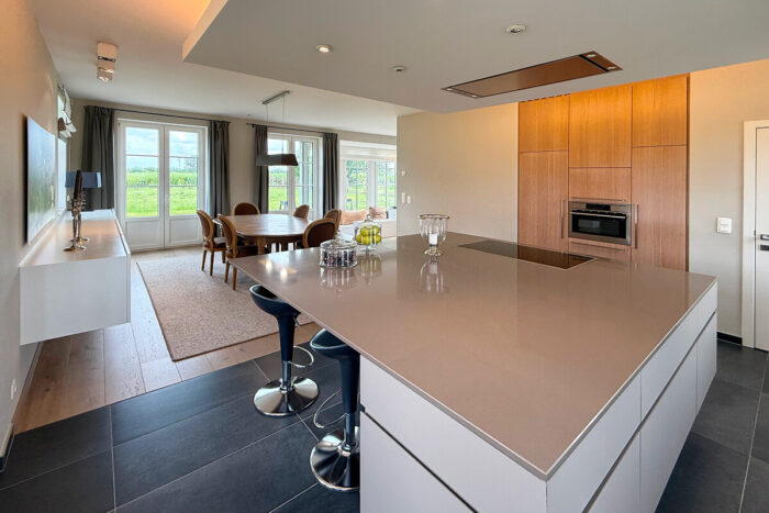 Moderne open keuken en eethoek in de kijkwoning Thorembais-Saint-Trond Blavier, met een groot eiland, strakke werkbladen, barstoelen, ingebouwde oven en houten kasten. De grote ramen zorgen voor natuurlijk licht en inspiratie huis.
