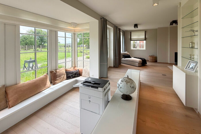 Moderne, lichte kamer met grote ramen en een zitje bij het raam met bruine kussens - perfect om inspiratie huis kijkwoning Blavier Thorembais-Saint-Trond- op te doen met houten vloeren, een wereldbol en een groen landschap dat buiten te zien is.