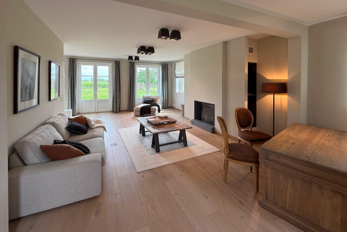 Een lichte, moderne woonkamer in de kijkwoning 1360 Perwez Blavier met lichte houten vloeren, beige sofa, open haard en grote ramen met uitzicht op groen. Neutrale tinten en veel natuurlijk licht zorgen voor een gezellige uitstraling.