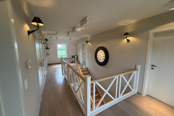 Lichte hal met houten vloeren, witte muren en een trap met witte leuning in kijkwoning Thorembais-Saint-Trond Blavier. Wandlampen voegen licht toe terwijl natuurlijk licht binnenstroomt vanuit een raam aan het einde, waardoor de grote klok boven de trap wordt benadrukt.