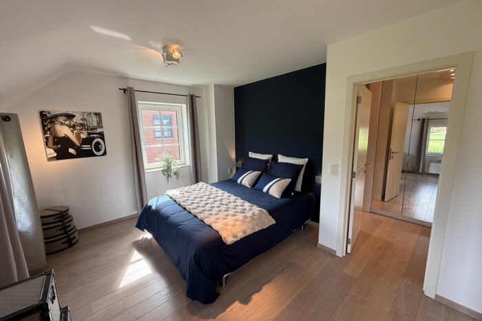 Een moderne slaapkamer in de kijkwoning Thorembais-Saint-Trond Blavier heeft een marineblauwe accentmuur, een tweepersoonsbed met blauw en wit beddengoed, twee ramen, een zwart-witfoto en een open deur naar een spiegelende hal vol natuurlijk licht.