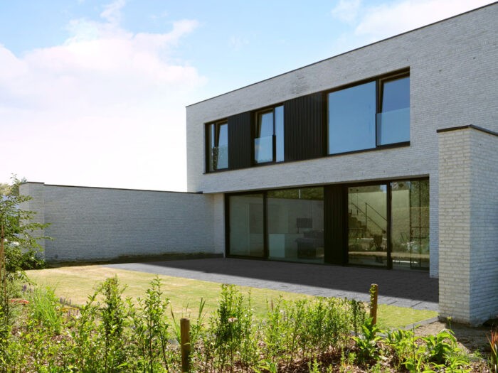 Maison moderne à deux étages avec de grandes fenêtres et un extérieur en briques claires. Le niveau inférieur est doté de portes vitrées et l'arrière-cour présente une pelouse simple avec un aménagement paysager clairsemé. Le ciel est clair avec quelques nuages.