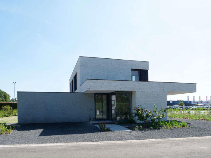 Une maison moderne et minimaliste à deux étages avec un toit plat, un extérieur gris clair et de grandes fenêtres. Elle dispose d'une entrée à cadre noir et est entourée d'une allée en gravier avec un aménagement paysager clairsemé. Le ciel est clair et bleu.