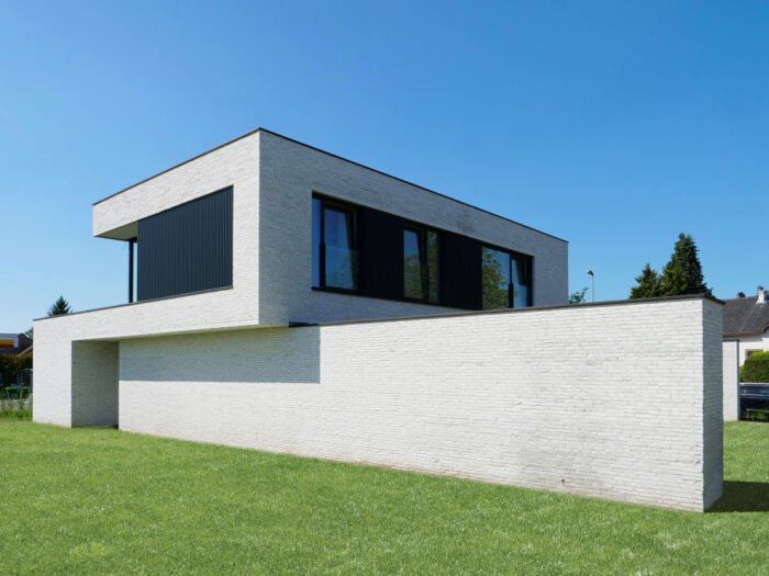Une maison moderne et minimaliste avec un toit plat et de grandes fenêtres à cadre noir. L'extérieur est revêtu de briques blanches et la structure est entourée d'une pelouse bien entretenue sous un ciel bleu clair.