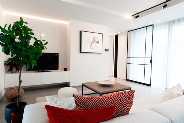 Un salon moderne avec un canapé blanc et des coussins à motifs rouges. Une télévision est installée sur un meuble bas blanc contre un mur clair, orné d'une œuvre d'art abstraite encadrée. Une plante en pot se trouve sur la gauche et de grandes fenêtres apportent de la lumière naturelle.