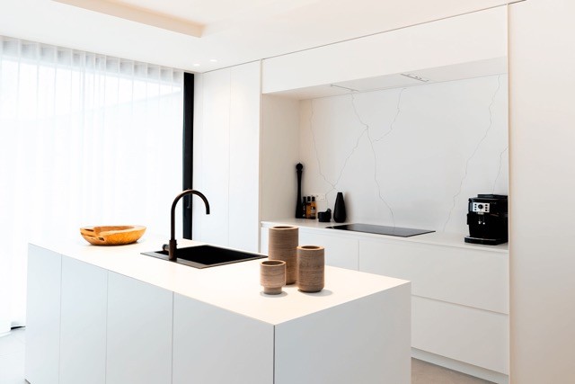 Une cuisine moderne et minimaliste avec des meubles blancs et un îlot. L'îlot est doté d'un évier et d'un robinet noirs, avec deux vases en céramique sur le dessus. Un bol en bois se trouve à proximité. Le mur du fond est doté d'appareils intégrés et de lignes décoratives subtiles.