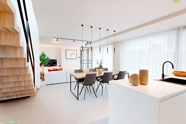 Un salon et une salle à manger modernes et ouverts au design minimaliste. Comprend une grande table à manger en bois avec des chaises noires, un îlot de cuisine élégant, un canapé blanc et des baies vitrées avec des rideaux transparents. Escalier en bois clair sur la gauche.