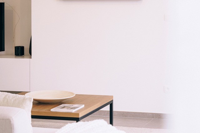 Un coin salon minimaliste avec une table basse en bois sur laquelle se trouve un plat rond en céramique et un magazine. Une partie d'un canapé blanc et d'une télévision sont partiellement visibles. La décoration est simple et moderne avec des tons neutres.
