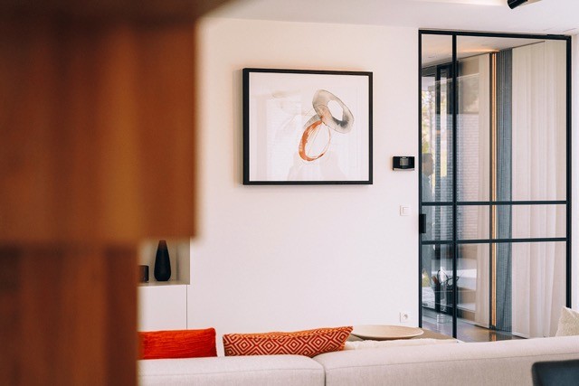 Un salon moderne avec un canapé blanc confortable avec des coussins rouges et à motifs. Une œuvre d'art abstraite encadrée est accrochée au mur. Une porte coulissante en verre mène à une autre pièce et la lumière naturelle filtre à travers, créant une ambiance chaleureuse.