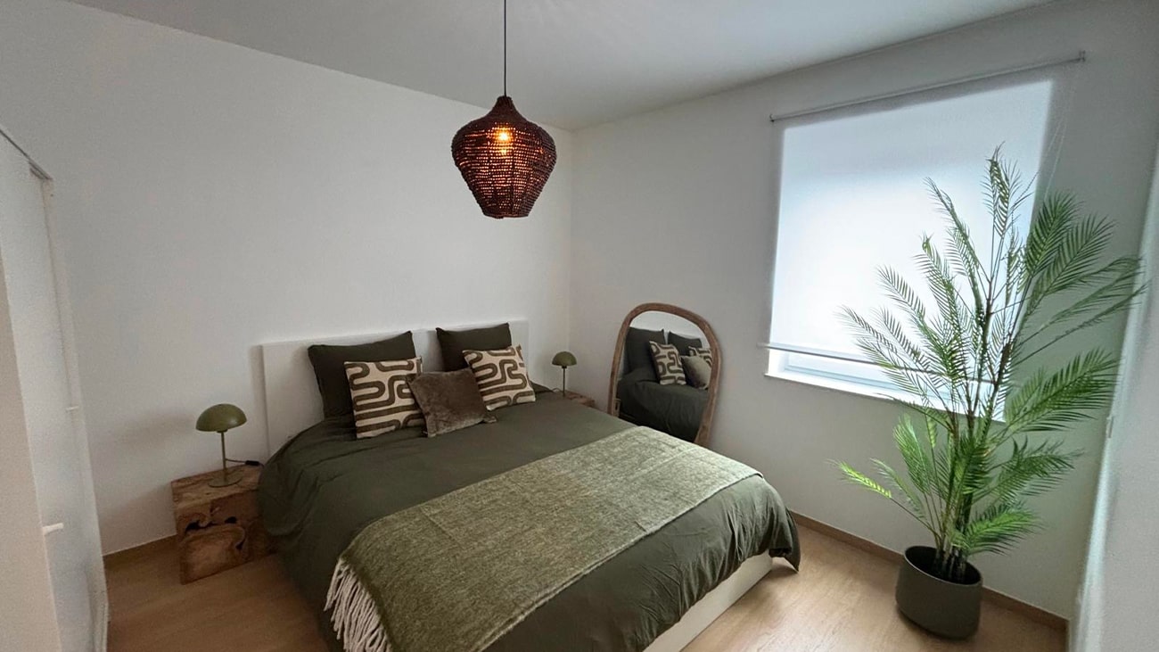 Une chambre minimaliste dans un appartement moderne de Jambes dispose dun lit double avec une literie vert foncé et deux oreillers à motifs. Une suspension en osier est suspendue au plafond, tandis quune grande plante en pot et un miroir rond sur un support en bois ornent le coin près de la fenêtre avec son store blanc.