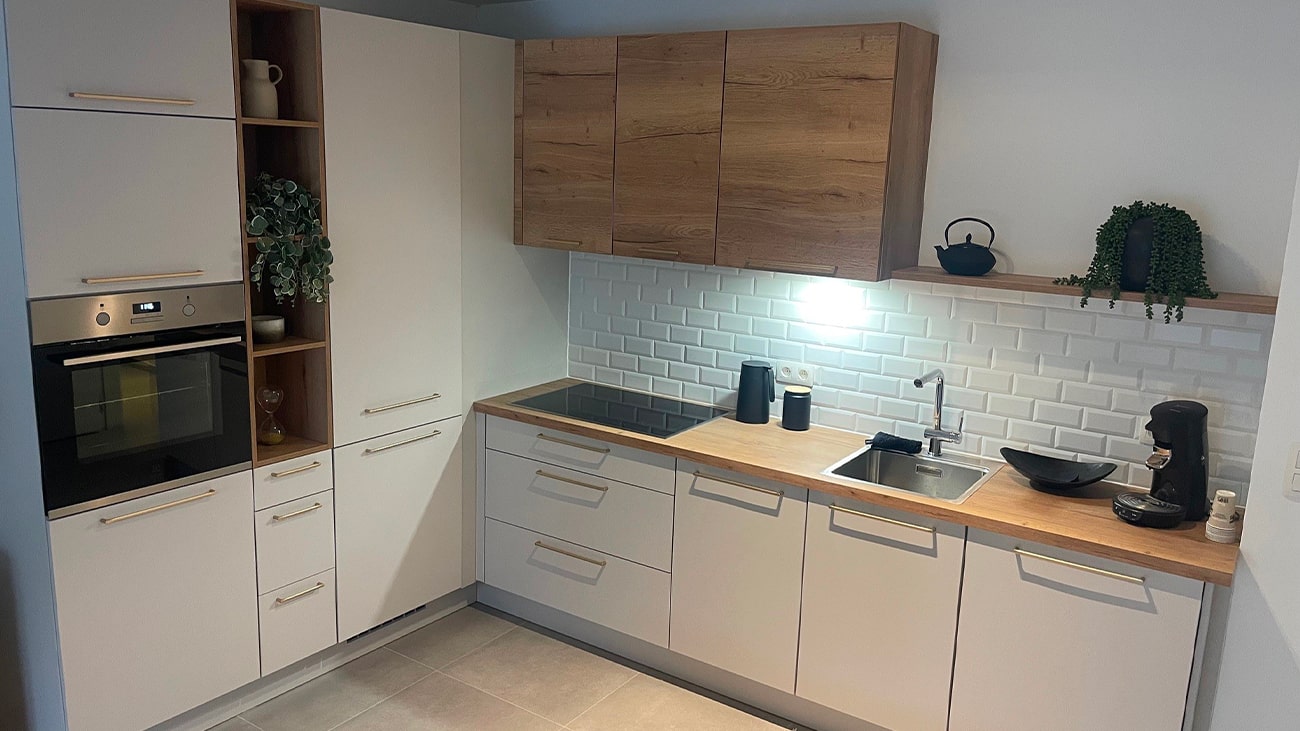 Cuisine moderne avec armoires blanches et touches de bois dans cet appartement moderne de Jambes. Comprend un four, une cuisinière, un évier et une cafetière. Une petite plante est posée sur une étagère au-dessus de lévier et une théière est posée sur le comptoir. Le dosseret en carreaux de métro ajoute un aspect épuré.