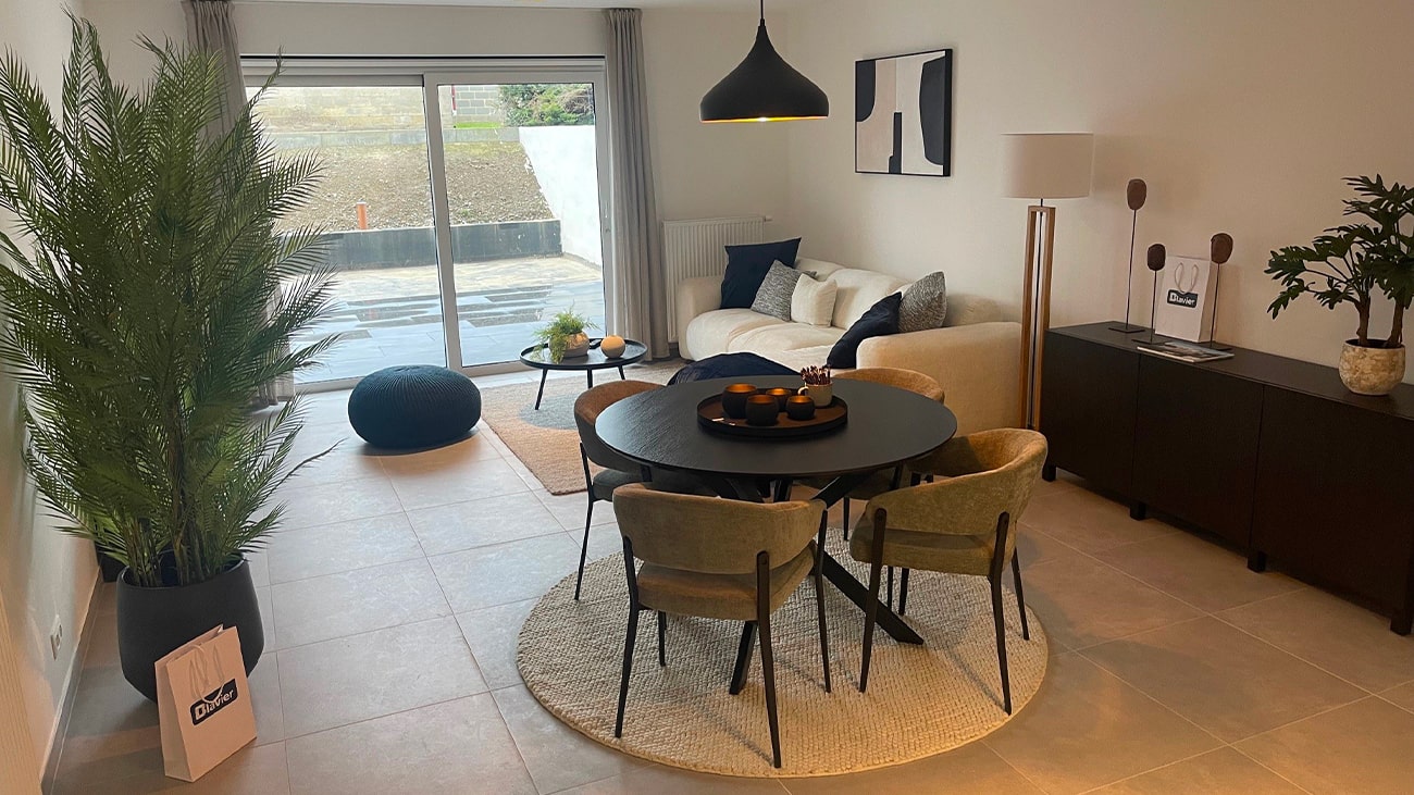 Salon moderne dans un appartement moderne à Jambes, avec une table à manger ronde noire et quatre chaises sur un tapis rond. Un canapé blanc avec des coussins foncés, une suspension noire et une grande plante rehaussent lespace. Des portes coulissantes en verre mènent à lextérieur, mettant en valeur un décor minimaliste et des tons neutres.