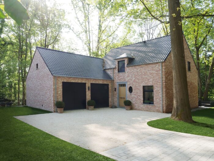 Een charmant bakstenen huis met een donker leien dak, omringd door hoge bomen. Het beschikt over een dubbele garage met zwarte deuren, een houten voordeur en potplanten bij de ingang. De oprit is van grind en leidt naar een goed onderhouden gazon.