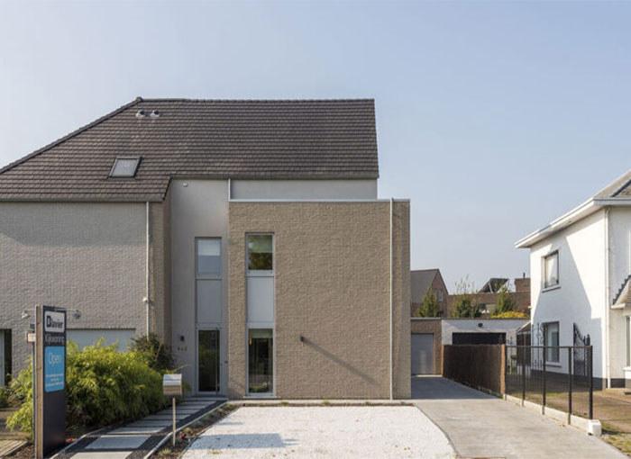 Maison moderne à deux étages avec une partie à toit plat et une structure principale à toit en pente. Façade en briques claires, grandes fenêtres et allée en gravier. Un panneau immobilier est visible à lavant. Maison attenante visible à droite. Pelouses et verdure clairsemées.