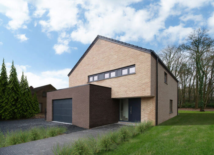 Une maison moderne en brique à deux étages avec un toit en pente et de grandes fenêtres. Le bâtiment dispose dune porte de garage sombre et dune entrée avant minimaliste. Il se dresse sur un ciel partiellement nuageux avec une pelouse et des arbres entourant la propriété.