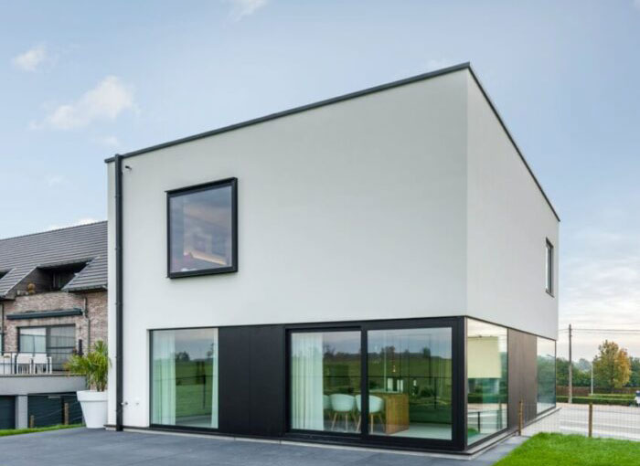 Maison moderne et minimaliste à deux étages avec une façade blanche et noire. De grandes baies vitrées laissent apparaître un coin repas. La maison dispose dun toit plat et est située sur une pelouse herbeuse sous un ciel nuageux.