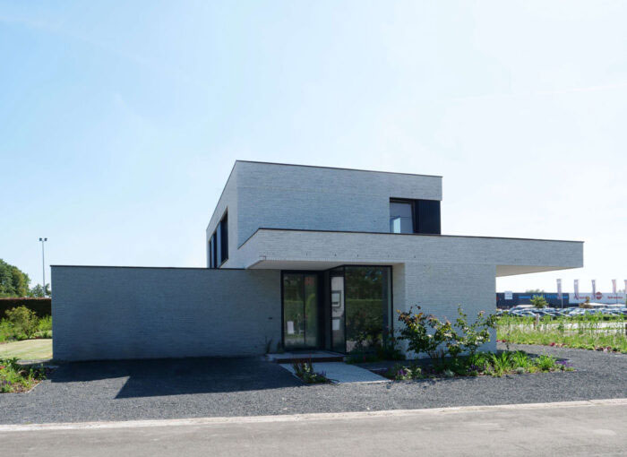 Maison moderne à deux étages au design minimaliste avec de grandes fenêtres et des toits plats. Le bâtiment est entouré dun chemin de gravier et dun aménagement paysager épuré, avec une pelouse ouverte et un ciel clair en arrière-plan.