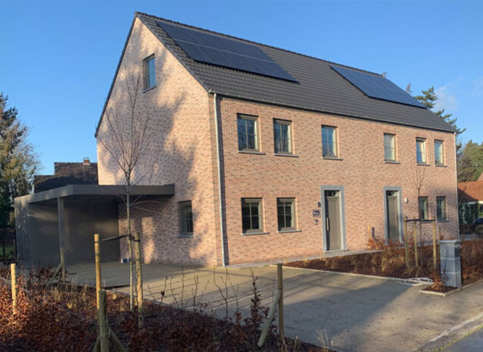 Une maison moderne en brique à deux étages avec un toit sombre et des panneaux solaires. Elle dispose de plusieurs fenêtres rectangulaires et dun abri pour voiture sur la gauche. Le jardin est récemment aménagé avec une clôture en bois et de jeunes arbres. Le ciel est clair et ensoleillé.