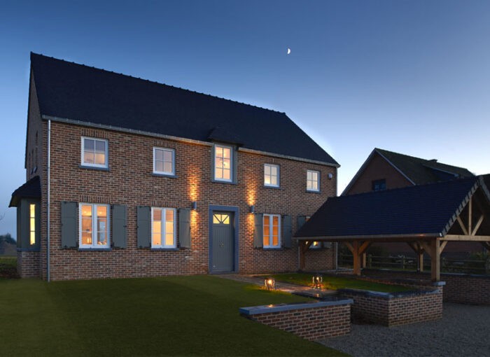 Een groot, twee verdiepingen tellend bakstenen huis met meerdere ramen is van binnenuit warm verlicht. Het heeft een donker dak en groene luiken. Naast het huis is een open garage of carport met een schuin dak. De lucht is helder met een halve maan.