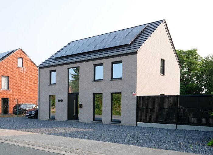 Maison moderne à deux étages avec façade en brique gris clair, avec panneaux solaires et grandes fenêtres. Adjacente à une clôture noire et une voiture garée, avec une autre maison en brique rouge à proximité. Une allée en gravier mène à l'entrée de cette Réalisation Maisons Blavier à Hautrage.