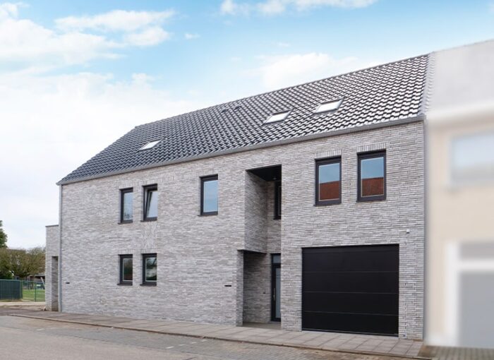 Maison moderne à deux étages avec un extérieur en briques grises et un toit en pente avec des tuiles. Elle possède de multiples fenêtres et une grande porte de garage noire. Le trottoir et la rue sont visibles au premier plan.