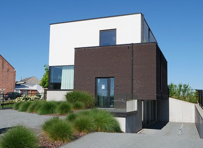 Een modern huis met twee verdiepingen en een minimalistisch ontwerp. De bovenste verdieping is wit, terwijl het onderste gedeelte donkere, getextureerde lambrisering heeft. Een pad leidt naar de ingang en siergrassen zijn langs de oprit geplant. Helderblauwe lucht op de achtergrond.