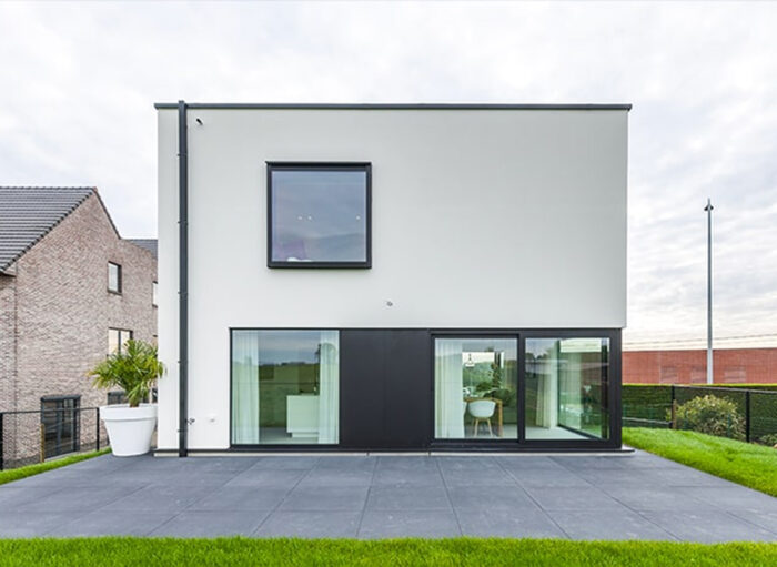 Modern kubusvormig huis met witte buitenkant, grote vierkante ramen en een plat dak. Op de begane grond zijn glazen schuifdeuren die naar een patio leiden. Links staat een grote plant in een witte pot. Het huis staat in een grasveld onder een bewolkte lucht.