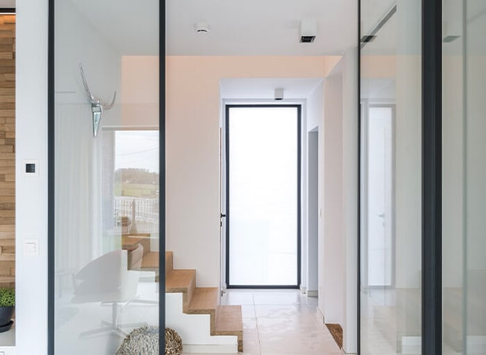 Minimalistische hal met grote glazen wanden en een centrale houten trap. Een hertengewei-decoratie is aan de muur bevestigd. Natuurlijk licht van een grote glazen deur verlicht de ruimte. Witte muren en lichte houten vloeren creëren een luchtige sfeer.