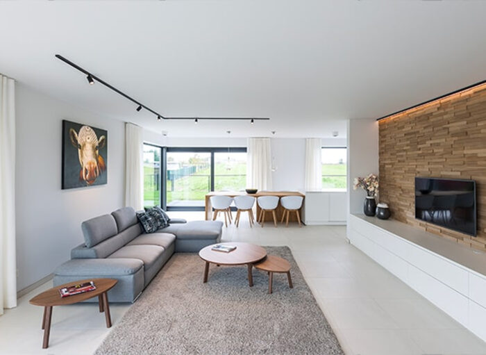 Moderne woonkamer met een grijze hoekbank, houten salontafels en een flatscreen-tv op een getextureerde accentmuur. Fel natuurlijk licht stroomt door grote ramen. Kunst aan de muur en een eettafel met stoelen op de achtergrond. Neutraal decorthema.