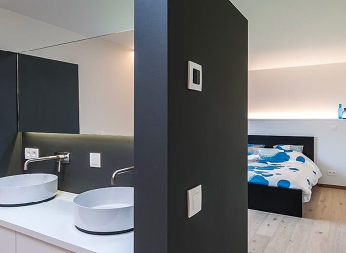 Modern interieur met een badkamer met twee ronde wastafels en een slaapkamer met een bed bedekt met blauw en wit beddengoed. Een scheidingswand scheidt de ruimtes, met een lichtschakelaar en thermostaat. De vloer is van hout en de kamer heeft een minimalistisch design.