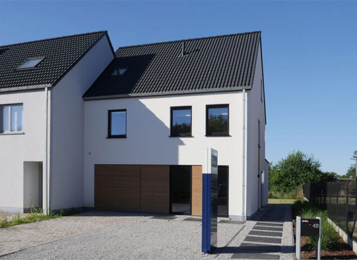 Maison moderne à deux étages avec un toit sombre en pente, un extérieur blanc et des panneaux en bois autour dun garage. Une allée pavée se trouve à lavant, menant à une entrée principale avec une porte vitrée à côté dun chemin paysager. Ciel clair et ensoleillé en arrière-plan.