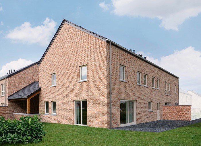 Une maison moderne en briques avec un toit en pente raide, avec plusieurs fenêtres et une porte coulissante en verre. Une petite parcelle dherbe et darbustes se trouve au premier plan, tandis que le ciel est partiellement nuageux.