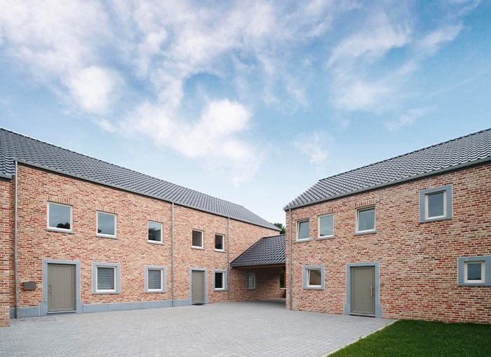 Deux bâtiments adjacents en briques avec portes et fenêtres grises, avec des toits en tuiles gris foncé. Les bâtiments forment une cour avec une surface pavée en pierre, sous un ciel bleu partiellement nuageux. Une ruelle étroite relie les deux structures.