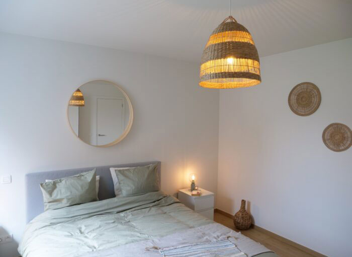 Une chambre minimaliste comprend un lit avec une literie vert clair, un miroir rond au-dessus de la tête de lit et deux décorations murales tissées. Une lampe suspendue tissée est suspendue au plafond et une petite lampe est posée sur une table de chevet blanche. Les murs sont blancs.
