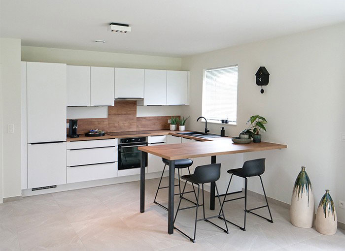 Cuisine moderne avec armoires blanches, comptoirs en bois et touches noires. Un bar à déjeuner avec deux tabourets noirs sétend depuis le comptoir. Une fenêtre fournit de la lumière naturelle et des vases décoratifs sont placés sur le sol. Une horloge murale est suspendue au-dessus.