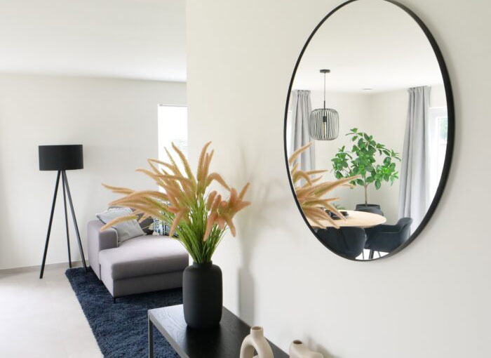 Un salon moderne avec un miroir rond reflétant une plante et une table à manger. Lespace comprend un canapé gris, un lampadaire noir, un tapis noir et un vase avec des plantes à plumes sur une console. Les tons neutres et la décoration minimaliste créent une atmosphère sereine.