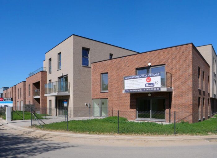 Immeuble d'appartements moderne aux façades en briques et aux tons clairs, avec de grandes fenêtres, des balcons et un panneau « À vendre » en français sur la façade. Un espace clôturé et un trottoir longent la rue en face.