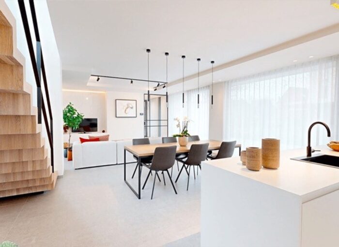 Moderne open woon- en eetkamer met een houten trap aan de linkerkant. Een eettafel met donkere stoelen staat in het midden onder hanglampen. Een witte bank staat tegenover een tv en grote ramen met doorschijnende gordijnen laten natuurlijk licht binnen. Minimalistisch decor.