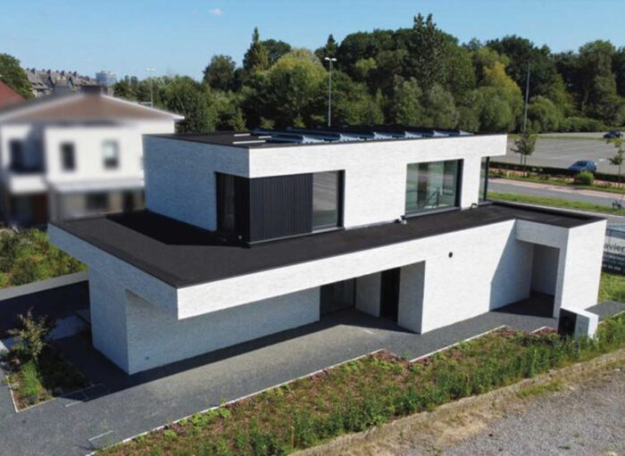 Modern, minimalistisch wit kubusvormig huis met plat zwart dak en grote ramen. Omgeven door een kleine tuin, staat het huis in een buitenwijk met bomen en een weg zichtbaar op de achtergrond.