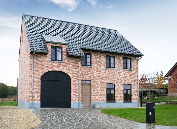 Une maison en brique à deux étages avec un toit en tuiles sombres. Elle est dotée de grandes fenêtres à cadre noir et dun mélange de portes de garage noires en bois et en arc. La façade est pavée de pavés et dune petite zone de gravier. Les environs sont gazonnés et arborés.