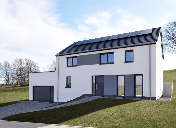 Maison blanche moderne à deux étages avec fenêtres sombres et toit en pente avec panneaux solaires. Un garage simple est attenant. La maison est située sur une pente herbeuse, avec un ciel clair et des arbres nus en arrière-plan.