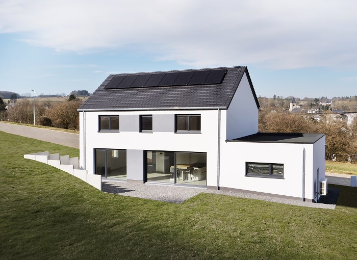 Maison moderne à deux étages avec un toit en pente et des panneaux solaires, de grandes portes vitrées et une entrée latérale. Entourée d'une pelouse verdoyante, la maison se dresse sous un ciel bleu parsemé de nuages, à côté d'une route.