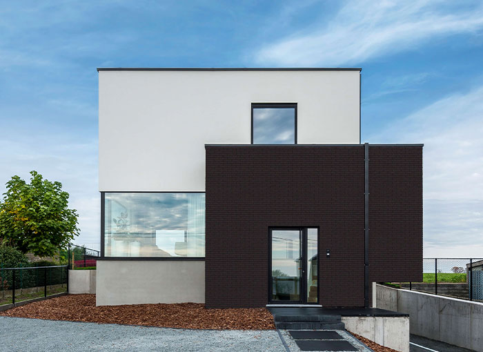 Maison moderne à deux étages au design minimaliste, avec des sections rectangulaires blanches et gris foncé, de grandes fenêtres et une cour avant en gravier bordée de paillis et d'un petit arbre sous un ciel partiellement nuageux.