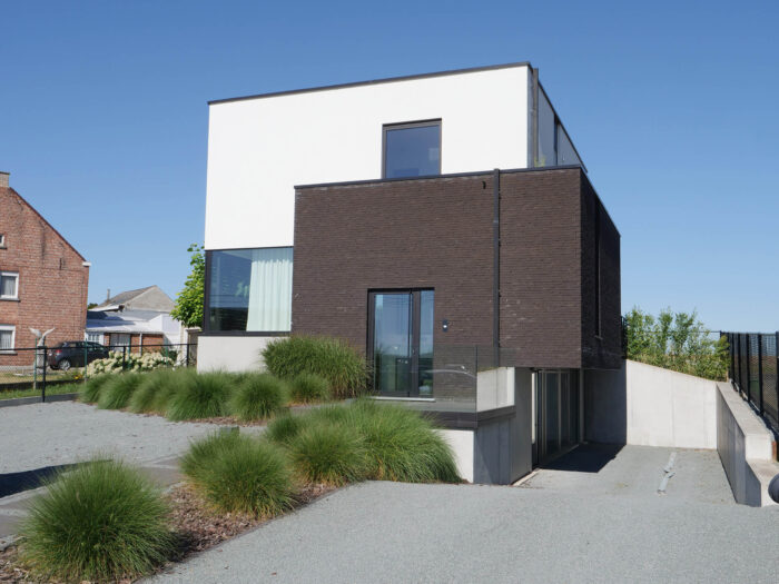 Een modern huis met twee verdiepingen, een witte bovenverdieping en een donkerbruine benedenverdieping, met grote ramen, glazen balustrades en omgeven door groene siergrassen.