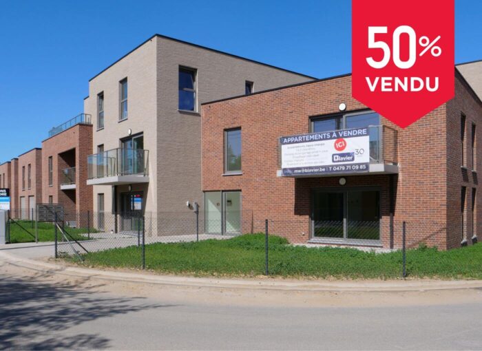 Immeuble d'appartements moderne avec façade en briques et beige, balcons en verre et panneau annonçant la vente d'appartements. Une grande bannière rouge dans un coin indique « 50 % Vendu », indiquant que la moitié des appartements sont vendus.