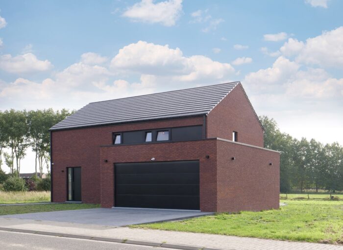Maison moderne à deux étages avec façade en briques rouges et toit à pignon foncé. Elle dispose dune grande porte de garage noire et de plusieurs fenêtres étroites. La maison est entourée dherbe et darbres, et le ciel est partiellement nuageux.